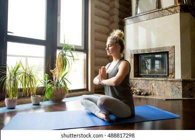 Full Length Portrait Of Attractive Young Woman Working Out At Home, Doing Yoga Exercise On Blue Mat, Sitting In Easy (Decent, Pleasant Posture) With Palms In Namaste, Meditating, Breathing, Relaxing