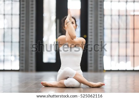 Full length portrait of attractive young woman working out in luxury fitness center, doing yoga or pilates exercise without mat on wooden floor. Gomukasana, Cow Face pose