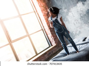 Full length portrait of beautiful Afro American girl in denim overall and headphones listening to music and smiling while standing on bed at home