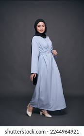 Full length portrait of a beautiful Asian female model wearing light blue dress with black hijab, isolated over dark background. Stylish Muslim female fashion lifestyle concept.