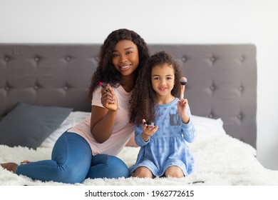 Full Length Portrait Of Beautiful Black Mother And Her Lovely Little Daughter Holding Makup Products And Smiling At Camera On Bed Indoors. Young Parent And Child Using Decorative Cosmetics At Home