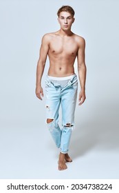Full length portrait of a beautiful blonde guy in light blue torn unbuttoned jeans without a shirt posing in the studio on a light blue background. Denim fashion. Men's beauty.