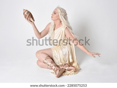 Full length portrait of beautiful blonde woman wearing a fantasy goddess toga costume with  magical crown.
Seated pose holding an ocean seashell, sitting on floor. isolated on white studio background.