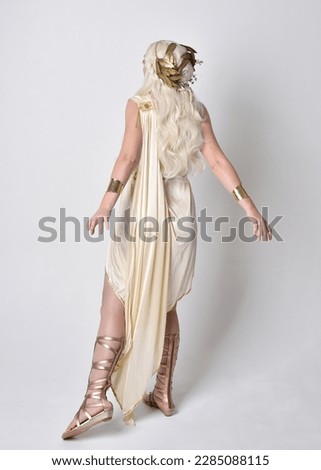 Full length portrait of beautiful blonde woman wearing a fantasy goddess toga costume with  magical crown.
Standing pose, facing backwards away from camera.  isolated on white studio background.