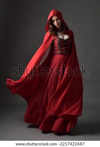 Full length portrait of beautiful brunette woman wearing red medieval fantasy costume with long skirt and flowing hooded cloak.
Standing pose with gestural hand poses, isolated on grey studio backgrou