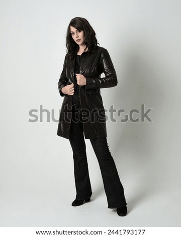 Full length portrait of beautiful brunette woman wearing long black leather trench coat, film noir detective. Standing pose, walking towards the camera, reaching out. Isolated white studio background