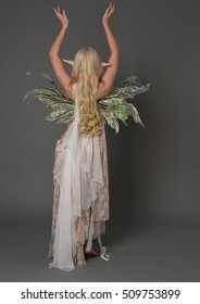 full length portrait of a beautiful fairy girl with long blonde hair, pointy ears and wings. seated pose against a grey background.