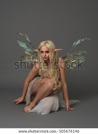 full length portrait of a beautiful fairy girl with  long blonde hair, pointy ears and wings.
seated pose against a grey background.
