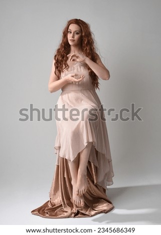 Full length portrait of beautiful  female model with long  brunette hair wearing a creamy pink gown dress. graceful sitting pose isolated on white studio background.