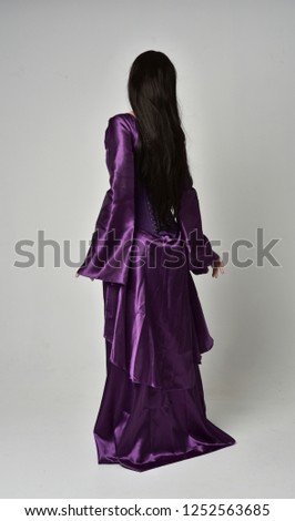 full length portrait of beautiful girl with long black hair,   wearing purple fantasy medieval gown. standing pose on grey studio background.