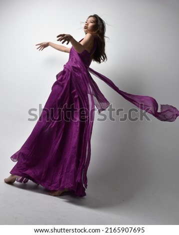 Full length portrait of beautiful girl wearing flowing fantasy purple ball gown. Standing with gestural hand poses, fro ma low angle perspective Isolated on studio background.