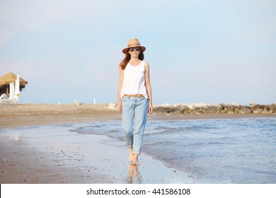 Full length portrait of beautiful middle aged woman wearing straw hat and sunglasses walking on the beach. 