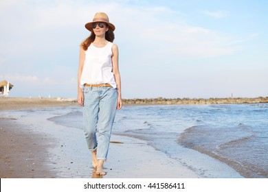 Full Length Portrait Of Beautiful Middle Aged Woman Wearing Straw Hat And Sunglasses Walking On The Beach. 