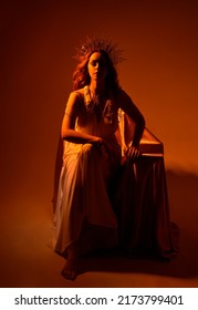Full length portrait of beautiful red head woman wearing long flowing fantasy toga gown with golden halo crown jewellery,  sitting pose  on a dark moody background with glowing or - Shutterstock ID 2173799401