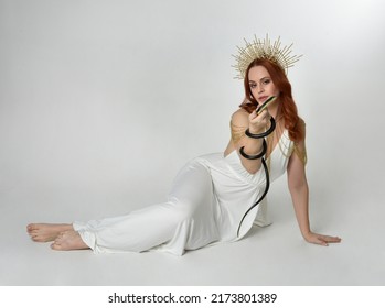 Full length portrait of beautiful red head woman wearing long flowing fantasy toga gown with golden halo crown jewellery,  sitting pose isolated on a white studio background.