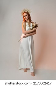 Full Length Portrait Of Beautiful Red Head Woman Wearing Long Flowing Fantasy Toga Gown With Golden Halo Crown Jewellery, Standing Pose   Isolated On A White Studio Background.