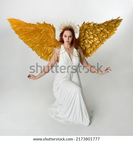 Full length portrait of beautiful red head woman wearing long flowing fantasy toga gown with golden halo crown and angel wings,  sitting pose isolated on a white studio background.