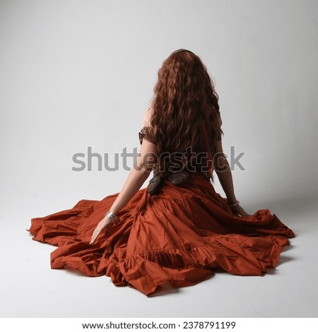 Full length portrait of beautiful red haired woman wearing a medieval maiden, fortune teller costume.  Kneeling pose, sitting down on floor. isolated on studio background.