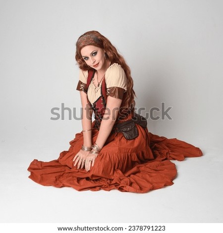 Full length portrait of beautiful red haired woman wearing a medieval maiden, fortune teller costume.  Kneeling pose, sitting down on floor. isolated on studio background.