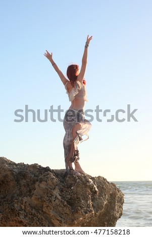 full length portrait of a beautiful red haired women, wearing shredded, cast away clothing. photographed at sunset with golden light beside the ocean.