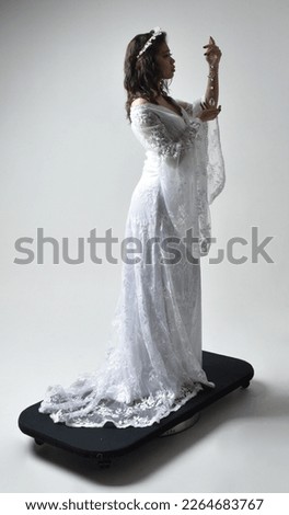 Full length portrait of beautiful woman wearing  fantasy costume, white bridal gown.  Backwards standing pose walking away,  gestural arm movements casting a spell. Isolated white studio background. 