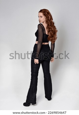 Full length portrait of beautiful woman with long red hair wearing black corset top and leather pants. Standing pose, facing backwards walking away from camera. Isolated on white studio background.