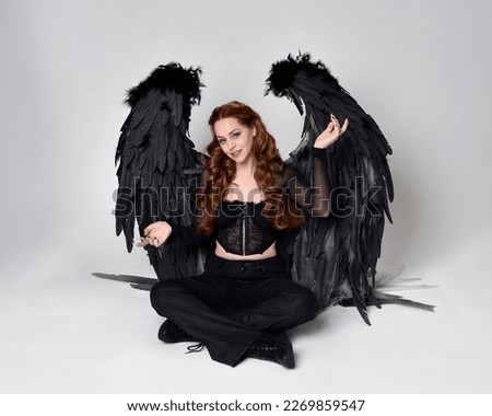 Full length portrait of beautiful woman with long red hair wearing sheer corset top, leather pants, large black angel feather wings.  Sitting pose with gestural hands reaching out, kneeling on floor.