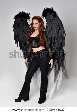 Full length portrait of beautiful woman with long red hair wearing sheer corset top, leather pants,  large black angel feather wings. Standing pose, walking forwards with gestural hands reaching out. 