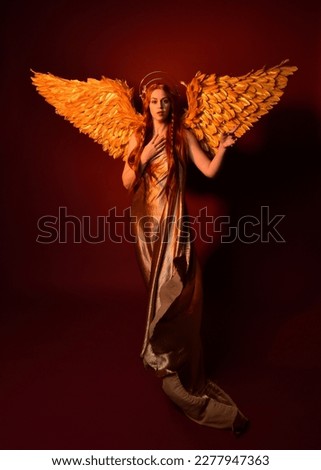 Full length portrait of beautiful woman model with long red hair, gold silk robes, crown  fantasy feather angel wings. Standing pose gestural hands reaching out isolated on dark red studio background