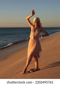 Full Length Portrait Of Beautiful Young Woman With Long Hair Wearing Flowing Dress, Standing Pose.  Ocean Beach Background With Back Lit Sunset Lighting.