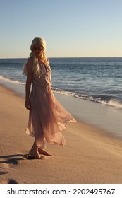 Full Length Portrait Of Beautiful Young Woman With Long Hair Wearing Flowing Dress, Standing Pose.  Ocean Beach Background With Back Lit Sunset Lighting.