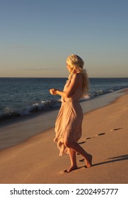 Full Length Portrait Of Beautiful Young Woman With Long Hair Wearing Flowing Dress, Standing Pose.  Ocean Beach Background With Back Lit Sunset Lighting.