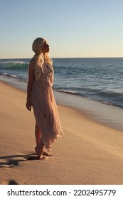 Full Length Portrait Of Beautiful Young Woman With Long Hair Wearing Flowing Dress, Standing Pose.  Ocean Beach Background With Back Lit Sunset Lighting.