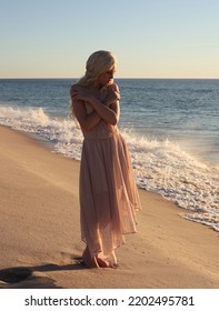 Full Length Portrait Of Beautiful Young Woman With Long Hair Wearing Flowing Dress, Standing Pose.  Ocean Beach Background With Back Lit Sunset Lighting.