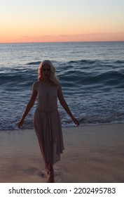 Full Length Portrait Of Beautiful Young Woman With Long Hair Wearing Flowing Dress, Standing Pose.  Ocean Beach Background With Back Lit Sunset Lighting.