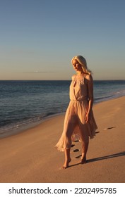 Full Length Portrait Of Beautiful Young Woman With Long Hair Wearing Flowing Dress, Standing Pose.  Ocean Beach Background With Back Lit Sunset Lighting.