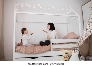 Full Length Portrait Of Beautiful Young Mother And Her Preschool Little Daughter In Pajamas Blowing Air Kisses To Each Other On Bed At Home. Cute Girl And Her Mom Enjoying Family Day.