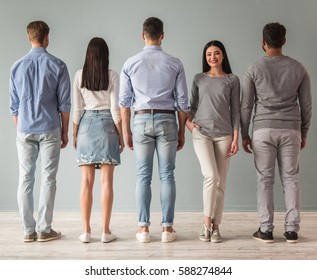 Full Length Portrait Of Beautiful Young People Standing In A Row, Their Backs Turned To Camera, One Girl Is Facing Camera And Smiling