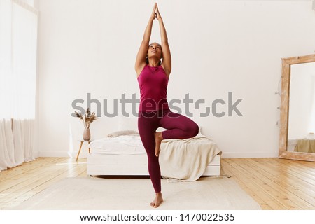Full length portrait of beautiful young Afro American woman with fit healthy body raising hands and placing sole of foot on her thigh, doing tree yoga stance or vriksasana in cozy bedroom interior