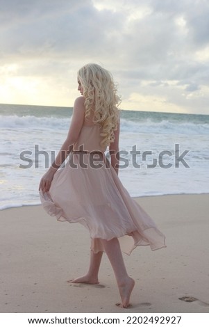 full length portrait of beautiful young woman with long hair wearing flowing dress, standing pose walking away from the camera.  ocean beach background with sunset lighting.