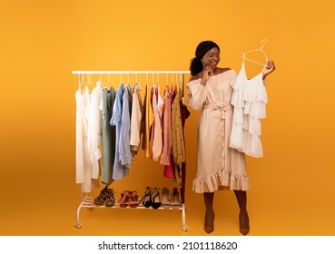 Full Length Portrait Of Black Female Stylist Holding Dress Near Clothing Rack, Choosing Trendy Outfit On Orange Studio Background. Shopping, Selecting New Wardrobe Concept