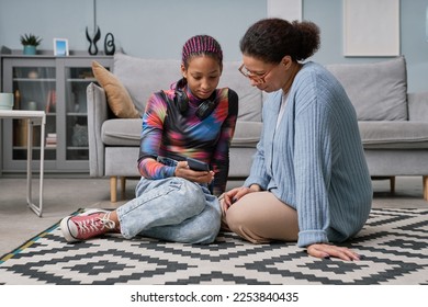 Full length portrait of black teenage girl talking to mom while sitting on floor at home and looking at smartphone screen - Powered by Shutterstock