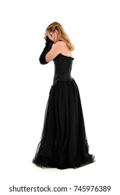 Full Length Portrait Of A Blonde Girl Wearing Black Gothic Gown, Standing Pose Isolated On White Background.