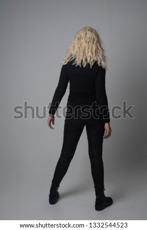 full length portrait of a blonde girl wearing  modern black jacket and pants, standing pose on grey studio background.