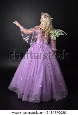 full length portrait of a blonde girl wearing a fantasy fairy inspired costume,  long purple ball gown with fairy wings,   standing pose with back to the camera on a dark studio background.