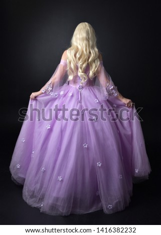 full length portrait of a blonde girl wearing a fantasy fairy inspired costume,  long purple ball gown,  standing pose with back to the camera on a dark studio background.
