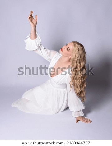 Full length portrait  of blonde woman  wearing  white historical bridal gown fantasy costume dress.  Sitting on floor posing with gestural hands reaching out, isolated on studio background.