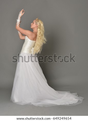 Full length portrait of a blonde woman wearing fur length  white gown and corset with a crown, standing against a grey studio background.