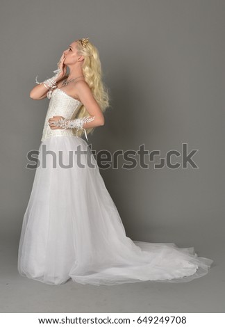 Full length portrait of a blonde woman wearing fur length  white gown and corset with a crown, standing against a grey studio background.