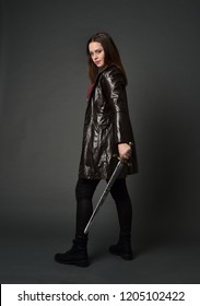 full length portrait of brunette girl wearing long leather coat and boots. standing pose holding a sword on a grey studio background